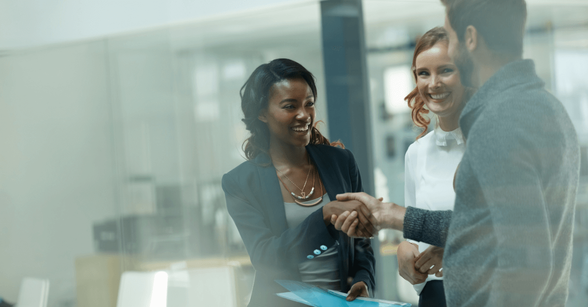 Image of three people standing in an office smiling at each other with two of them shaking hands