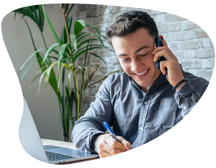 A man in a blue shirt who appears to be on a phone call, taking notes whilst sat at his desk. On the desk is also an open laptop.