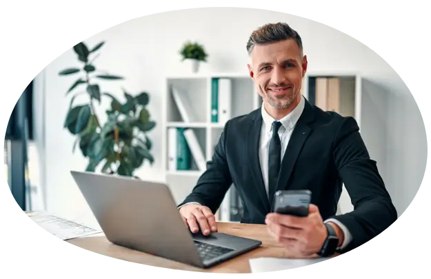 Professional man in a suit smiling with a cellphone in his left hand sitting behind a desk in an office. With an open laptop in front of him.